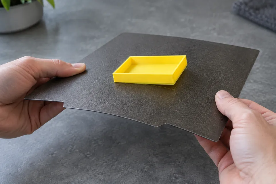 Hands gently flexing a removable textured steel sheet beneath a yellow printed tray.
